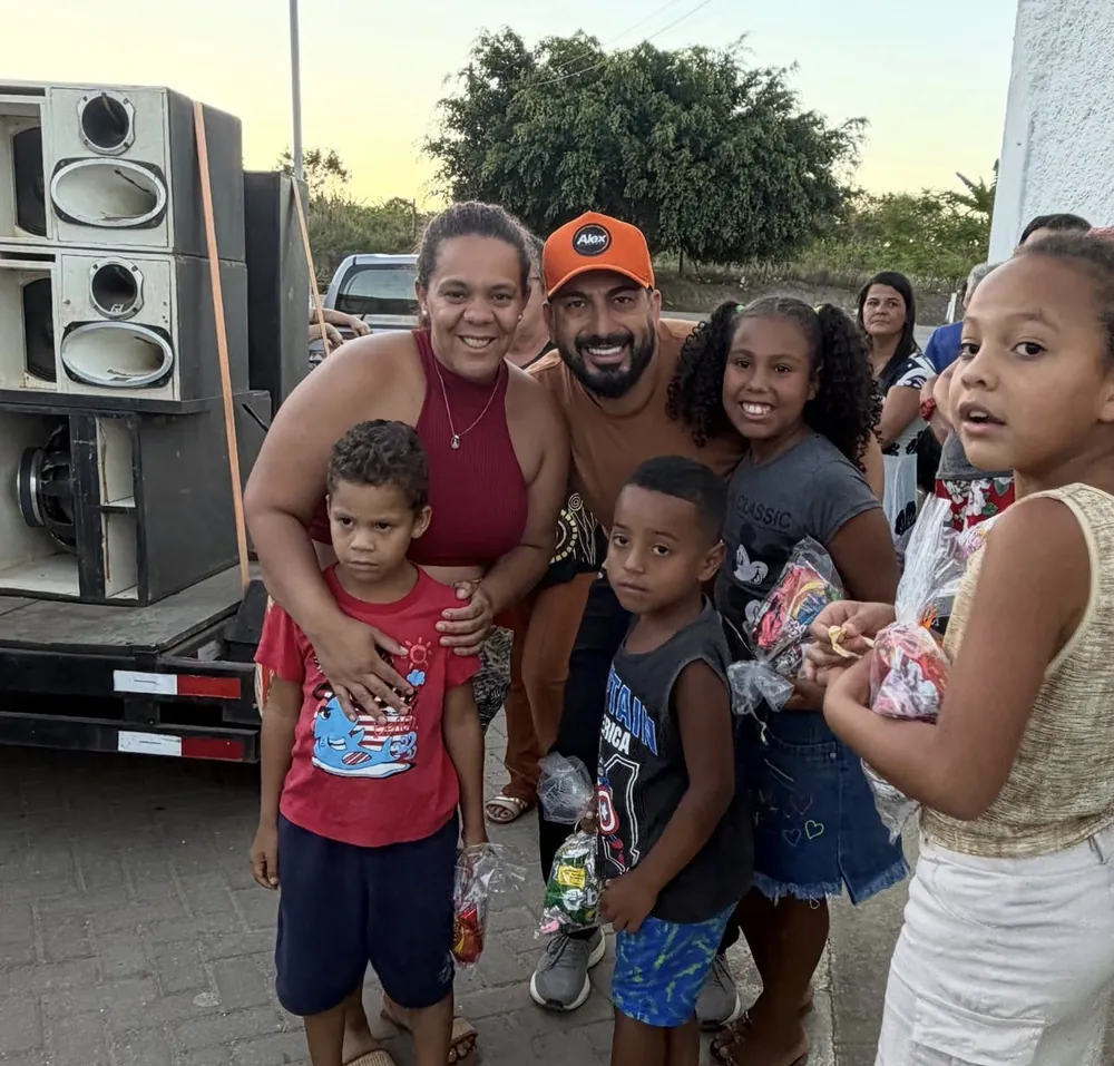 Alex sorrindo ao lado de uma mulher e de crianças que seguram sacolinhas de doces, perto de um carro de som