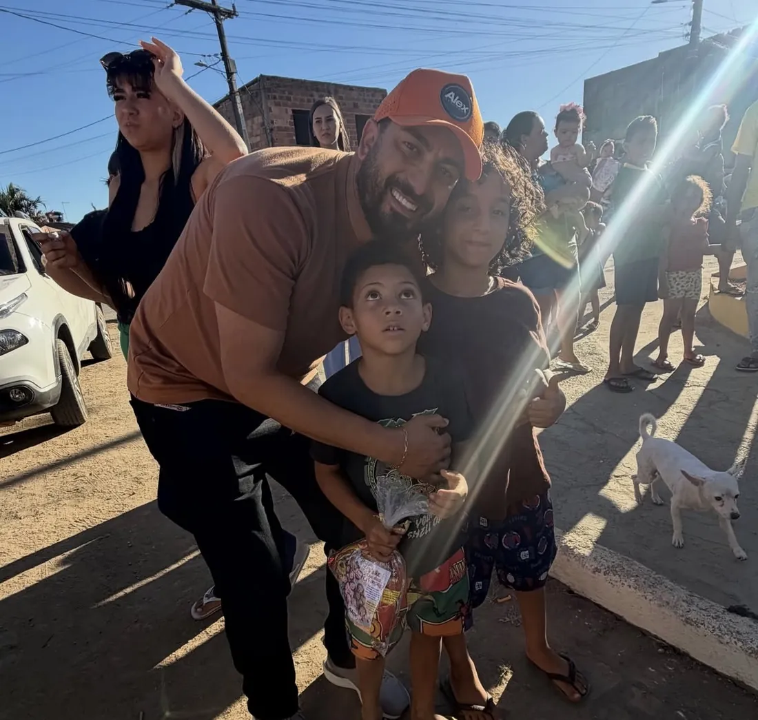 Alex abraçando duas crianças em uma rua de terra, com a luz do sol atravessando a imagem